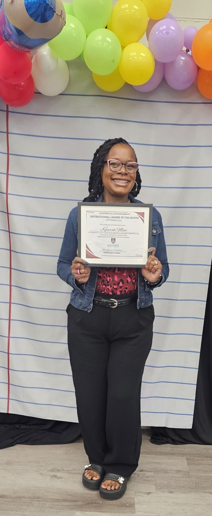 Keturah Mace wearing glasses, a denim jacket, and black pants, smiling warmly as she holds a framed Montgomery Public Schools "Instructional Leader of the Month" certificate. She stands in front of a backdrop styled like lined notebook paper with a arch of colorful balloons overhead.