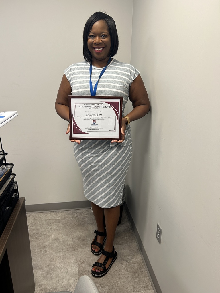 Amber Scott standing in an office setting, smiling and wearing a grey and white striped dress. She is holding a framed Montgomery Public Schools "Instructional Leader of the Month" certificate awarded for Academics & Accountability.