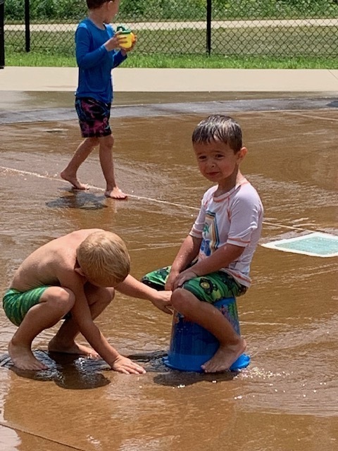 two elementary students at splash pad, one is sitting on a bucket