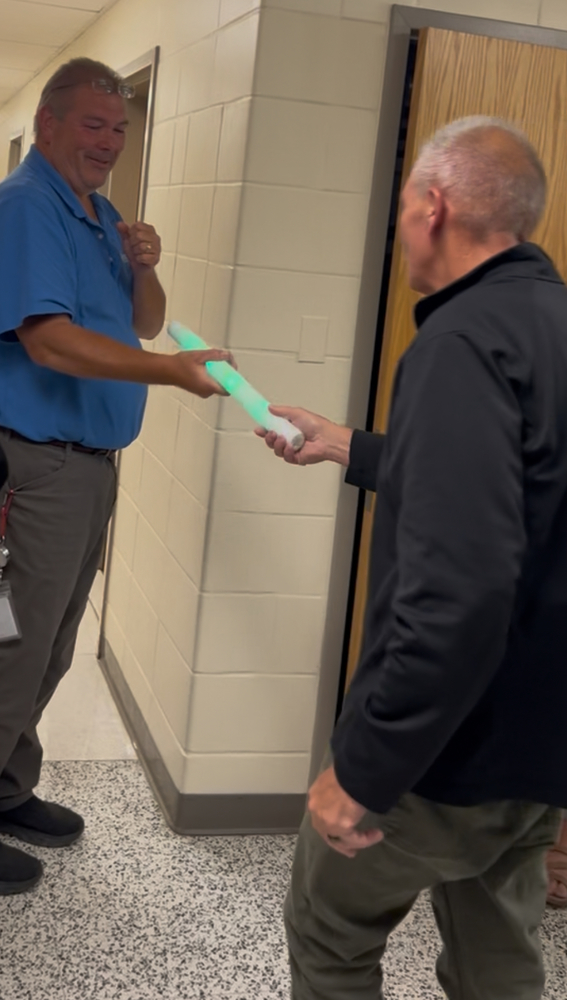 Two men stand in a hallway. One hands a glowing green toy sword to the other, both smiling. The atmosphere is lighthearted and playful.