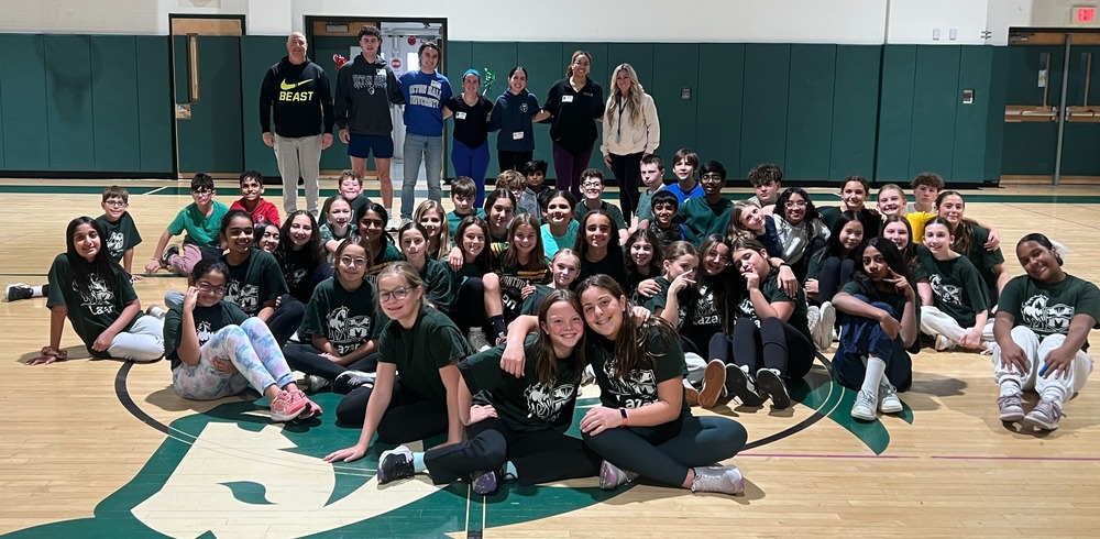 A large group of smiling children and adults pose in a gymnasium. The kids, wearing matching shirts, sit on the floor, while adults stand behind. The atmosphere is joyful. Basketball hoops are visible on each side.
