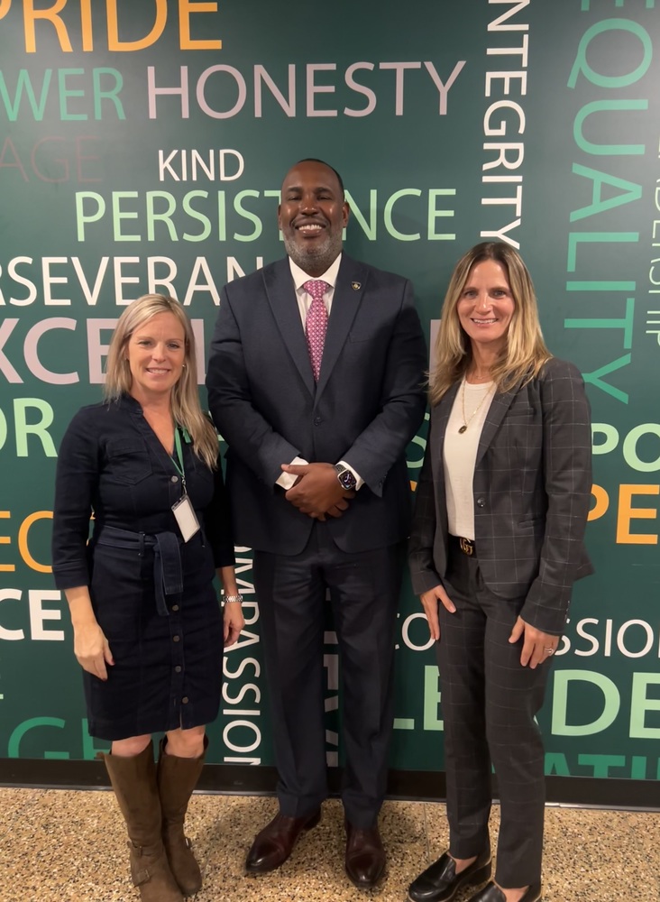 Three people stand smiling in front of a colorful word-filled wall with words like "HONESTY" and "KIND." The tone is positive and professional.