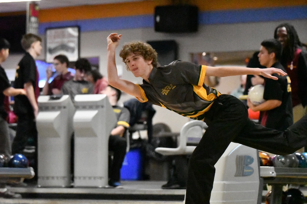 A young bowler in a bowling alley releases a ball with a focused expression, his arm extended. Teammates and spectators in the background add to the competitive atmosphere.