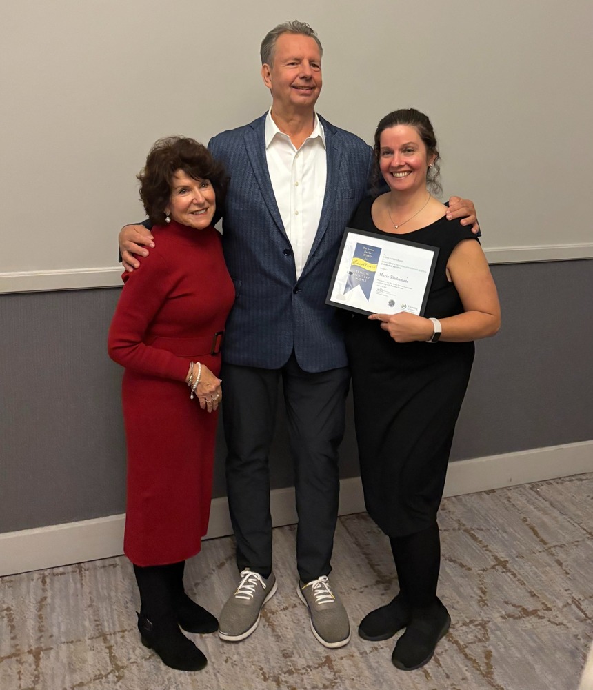 A woman in red, a man in a blue suit, and a woman in black holding a certificate stand smiling, conveying a joyful and celebratory atmosphere.