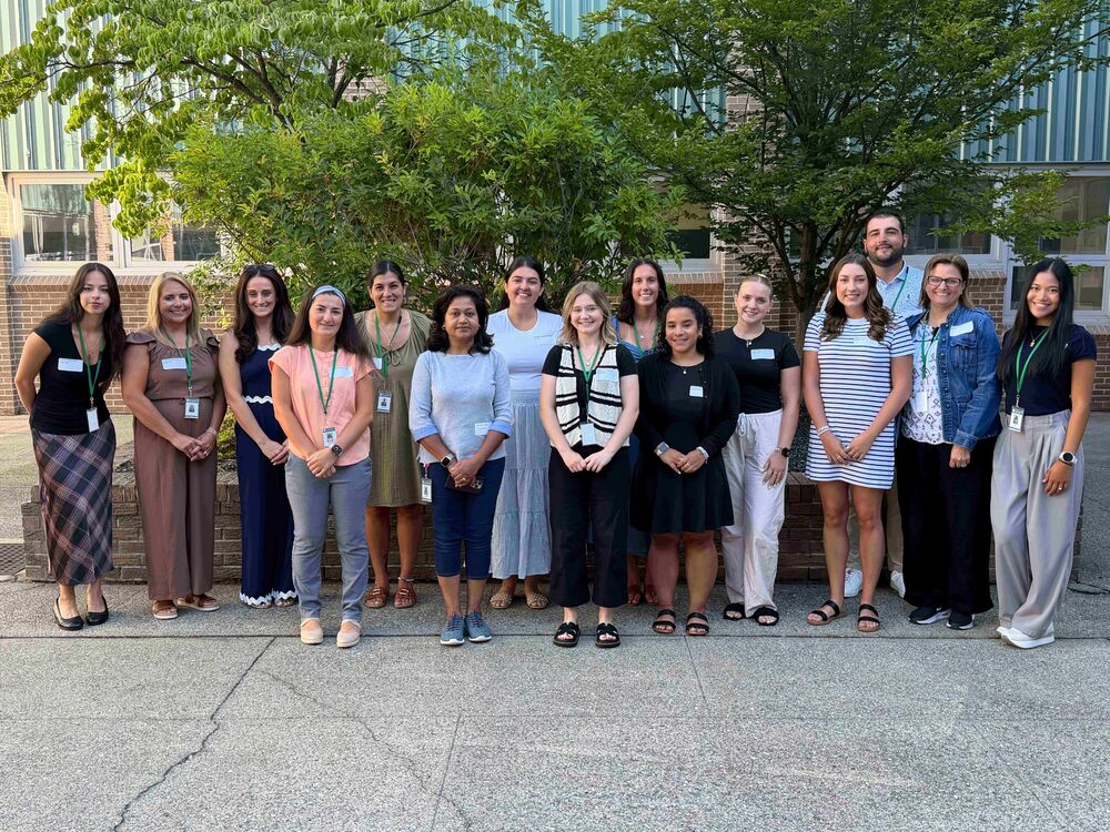15 new teachers standing in front of a large brick planter and two large trees.