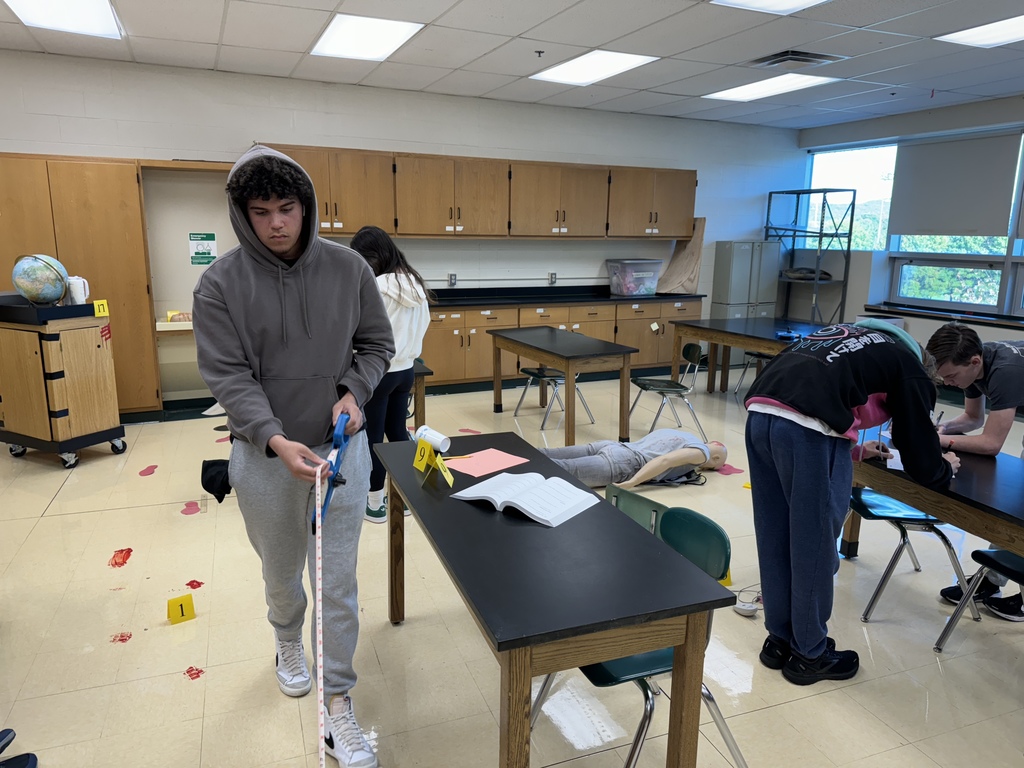 student taking measurements with a long measuring tape, evidence flags, red footprints and a model dummy facedown are also in the picture