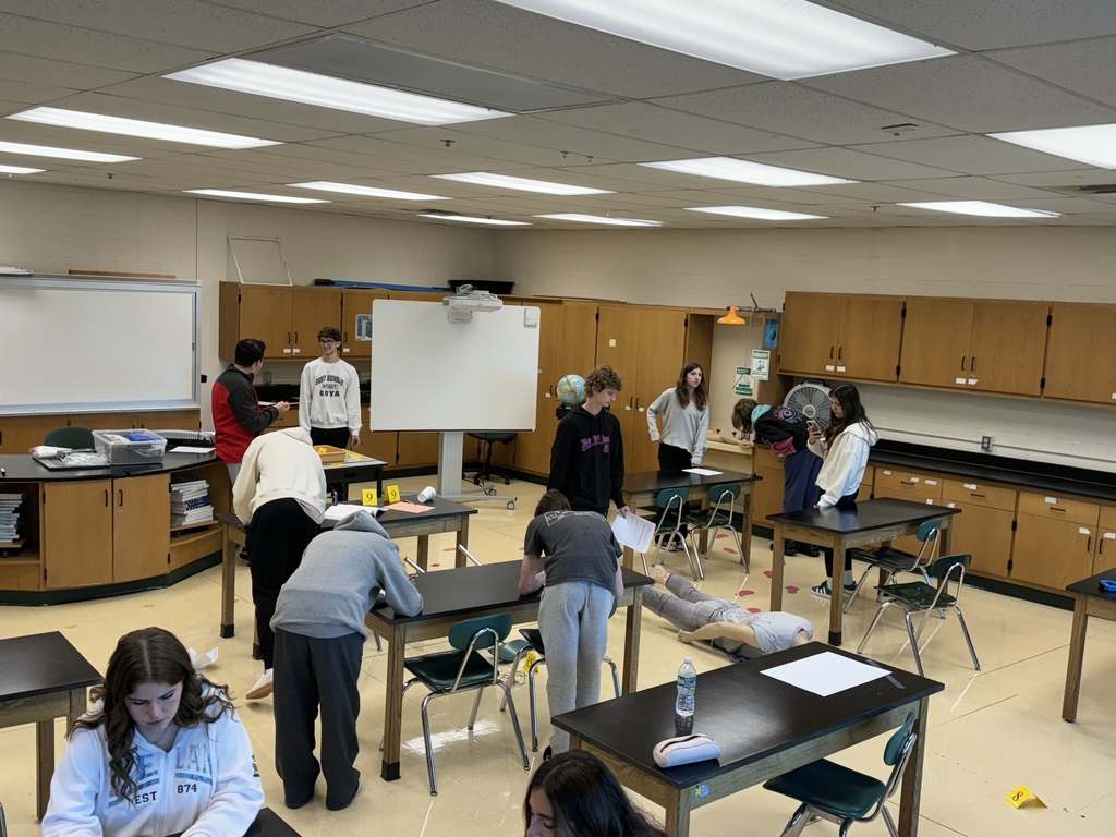 photo taken from a high angle of students at various lab tables working on their crime scene activity. Photo includes yellow evidence flags and a model dummy that is positioned facedown.