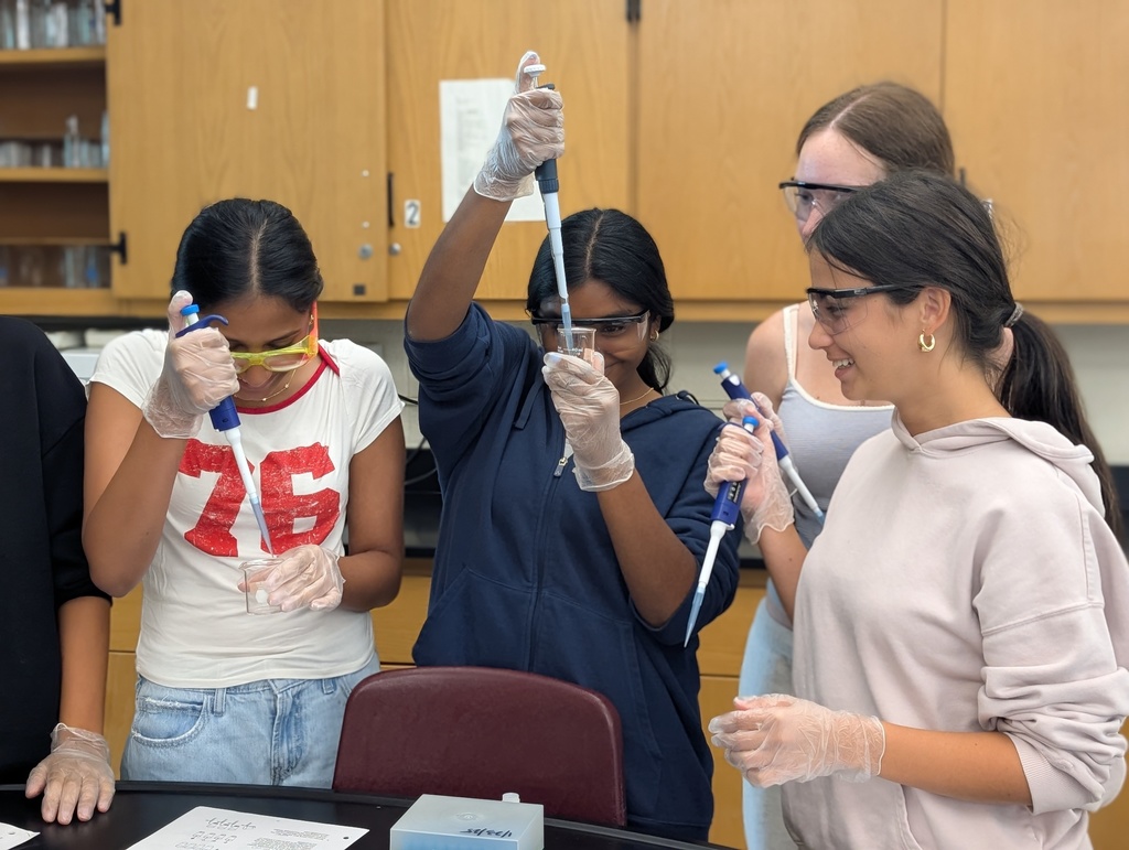 Five students sit at a black lab table wearing their saftey goggles. They are holding pipettes and extracting the colored dye from Skittles and M&M candies