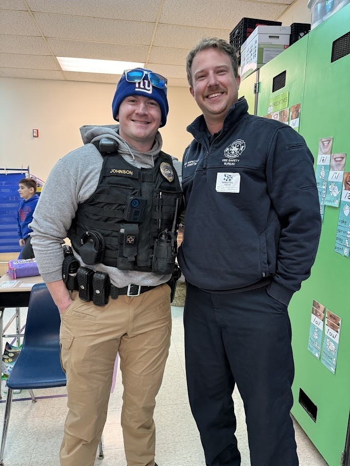 Two smiling men in a classroom. One is a police officer in uniform with a vest and beanie; the other wears a fire safety jacket. They stand closely together, showing camaraderie.