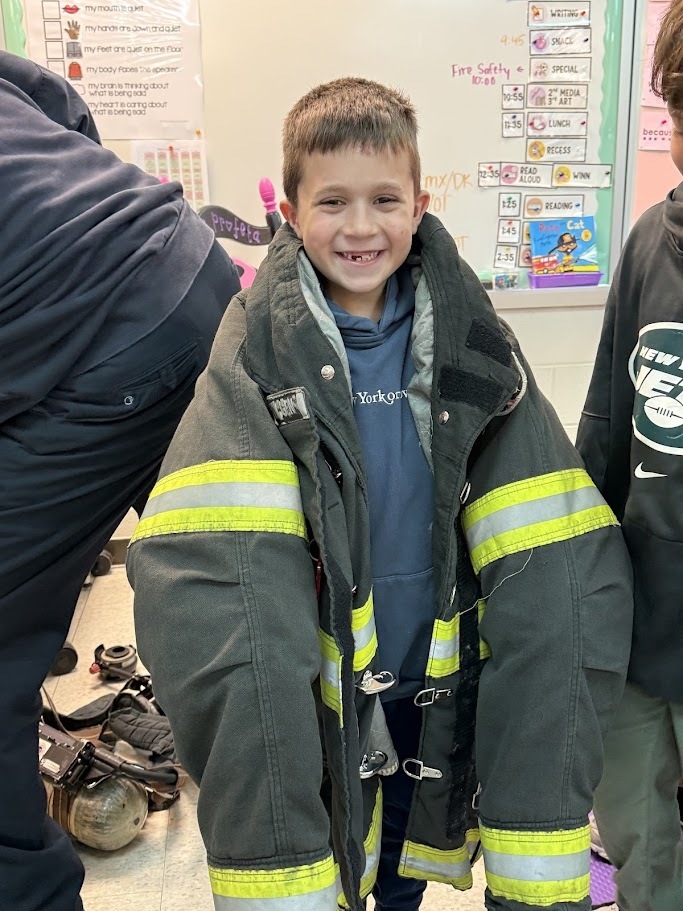 Young boy smiling while wearing an oversized firefighter coat. Classroom background with charts and a whiteboard. Playful and cheerful mood.