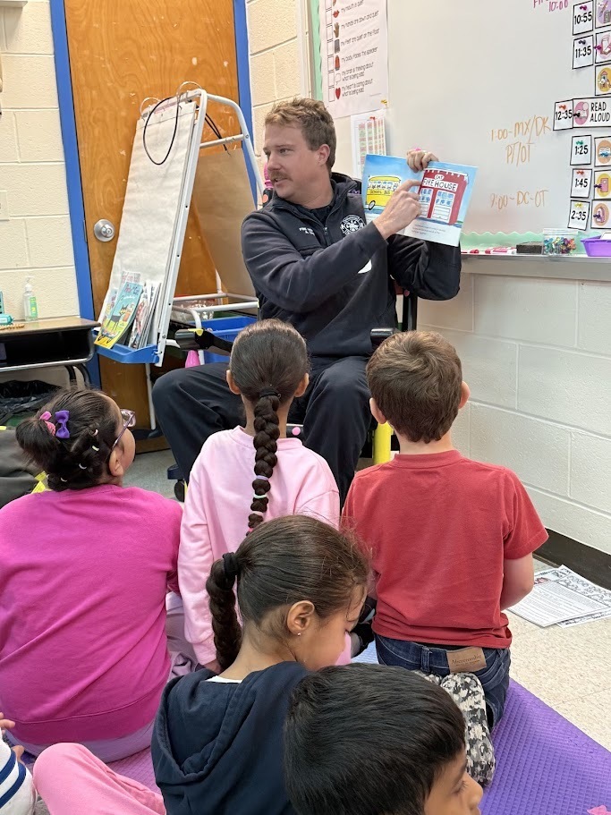 A man is reading a book aloud to young children seated on the floor in a classroom. The children look engaged, and the atmosphere is attentive and educational.