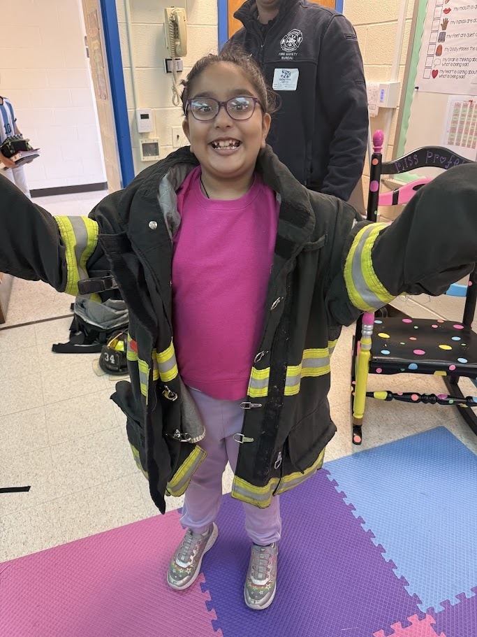 A smiling child stands with arms outstretched, wearing an oversized firefighter's jacket with yellow stripes. The setting is a classroom with colorful mats.