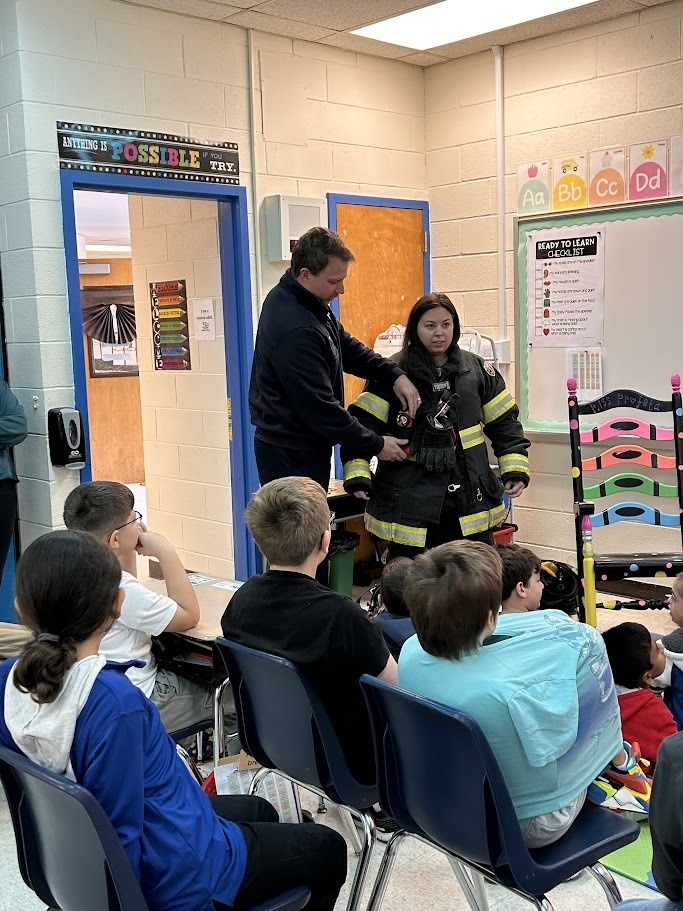 Firefighter demonstrates gear to attentive children in a classroom. The firefighter wears a black suit with yellow stripes, conveying a learning atmosphere.