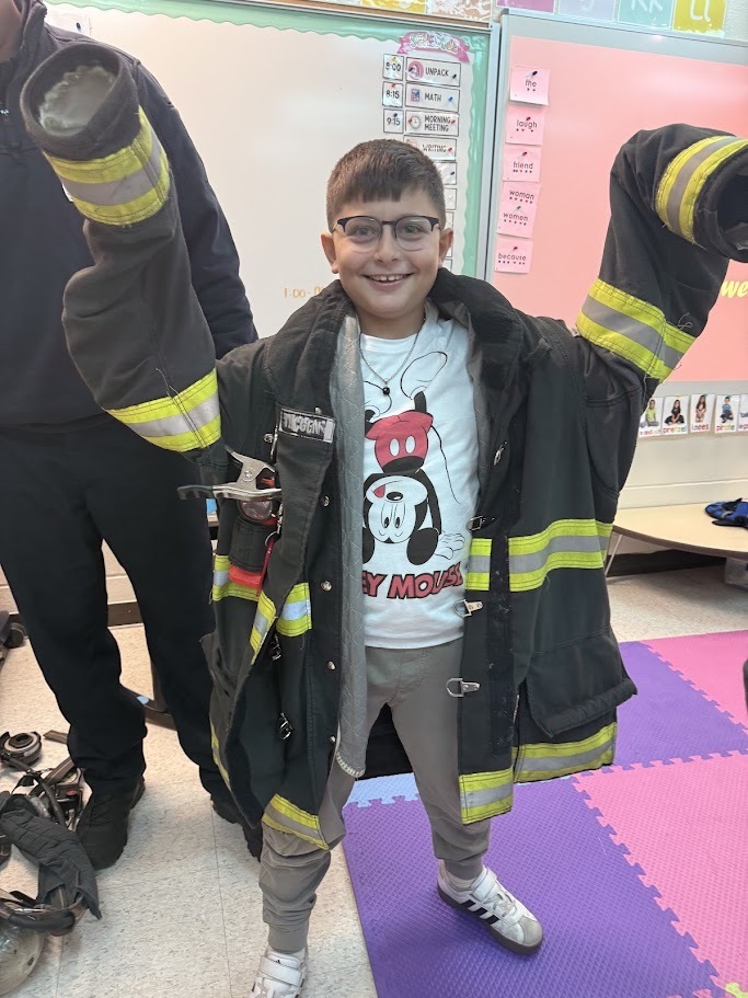 Young boy smiling and wearing an oversized firefighter jacket, sleeves extended, inside a colorful classroom with educational posters on the wall.