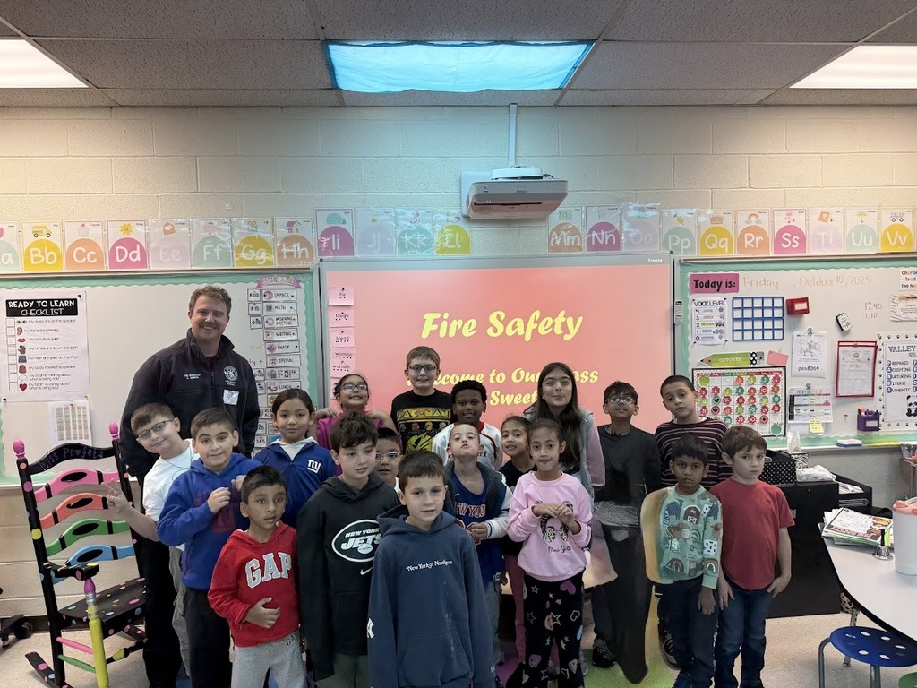 A group of children and an adult standing in a classroom in front of a screen displaying "Fire Safety." The children are smiling, and the room is colorful, creating a cheerful atmosphere.