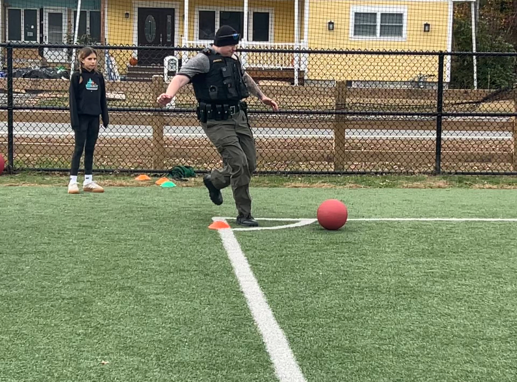 A police officer in uniform kicks a red kickball on a grassy field. A young girl watches nearby, with a fence and yellow house in the background.