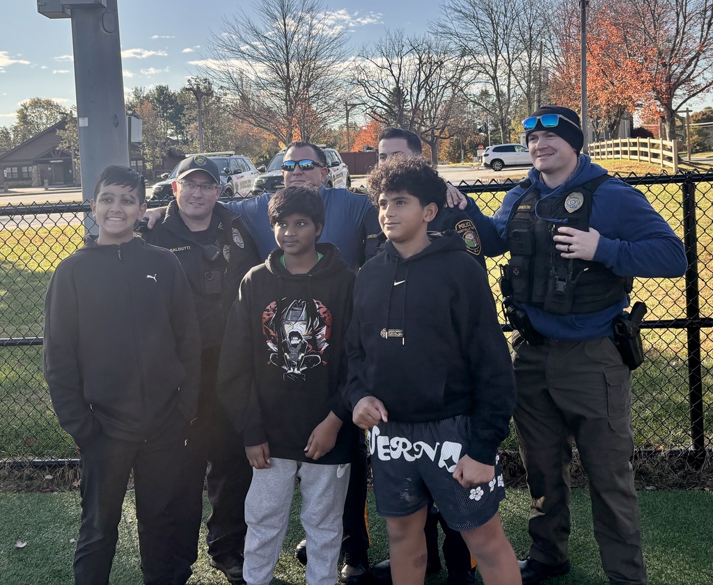 Three boys and four police officers pose and smile together in front of a chain-link fence at a park, with autumn trees in the background.