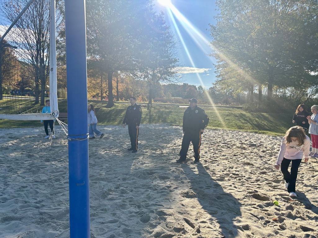 Children and two uniformed officers stand on a sunny sand volleyball court, surrounded by autumn trees. The atmosphere is playful and friendly.