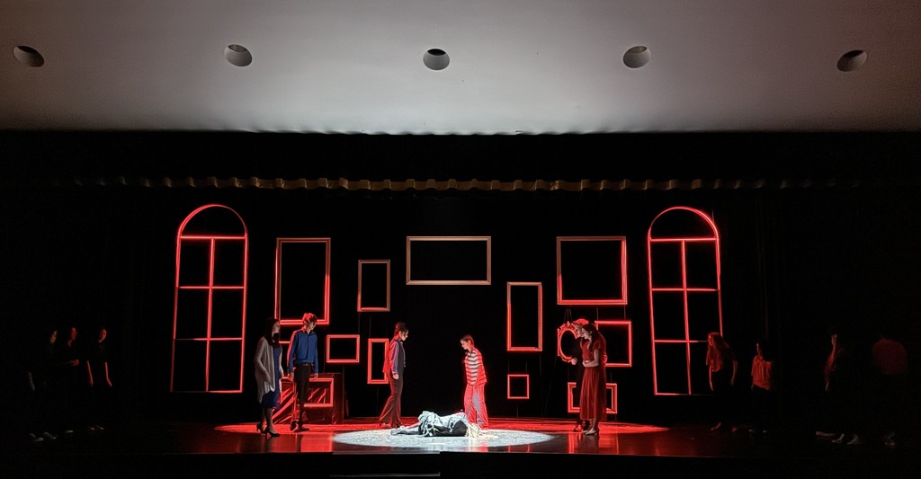 Theater stage with dim lighting shows a dramatic scene. Actors in red and blue stand around a person lying on the floor, framed by red-lit windows.