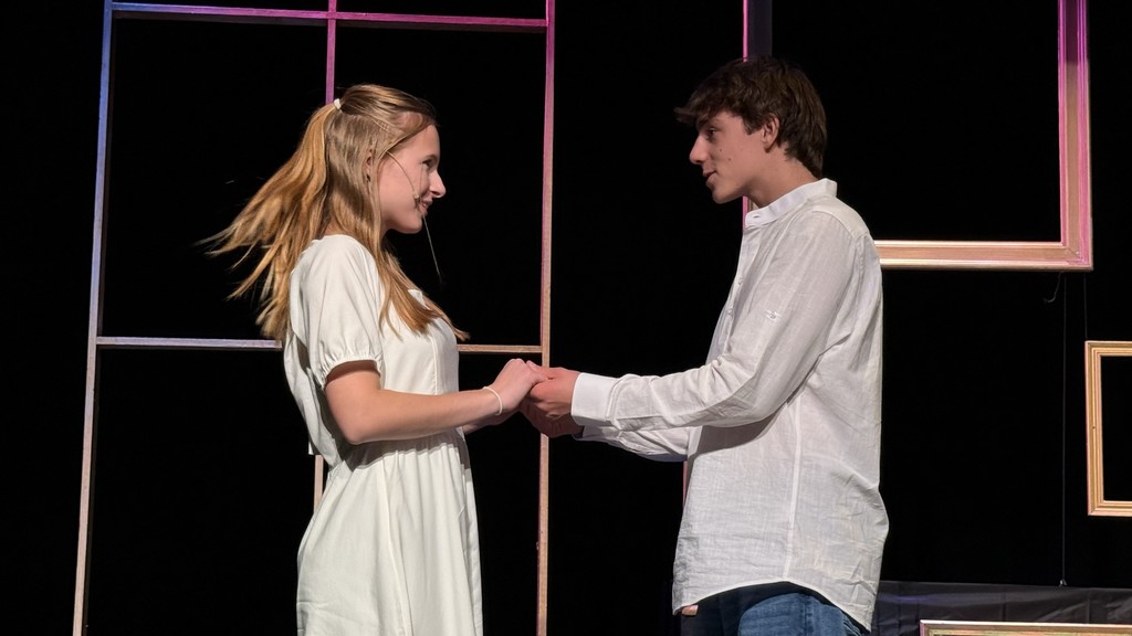 A young woman and man stand on stage, holding hands and looking at each other, conveying a tender moment. They are framed by geometric set pieces.