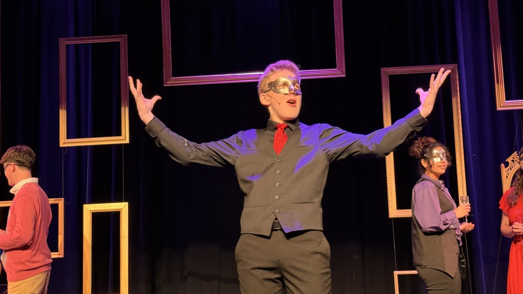 A person in a vest, red tie, and mask gestures dramatically on stage, surrounded by others in masks. Wooden frames hang in the background, creating a theatrical atmosphere.