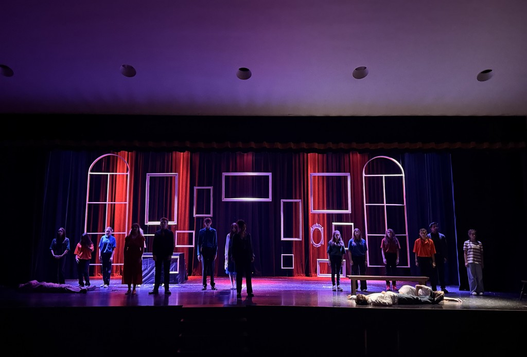 A theatrical performance on stage features actors standing solemnly in a line, with minimalist red and black geometric set designs and a person lying downstage, conveying drama and tension.