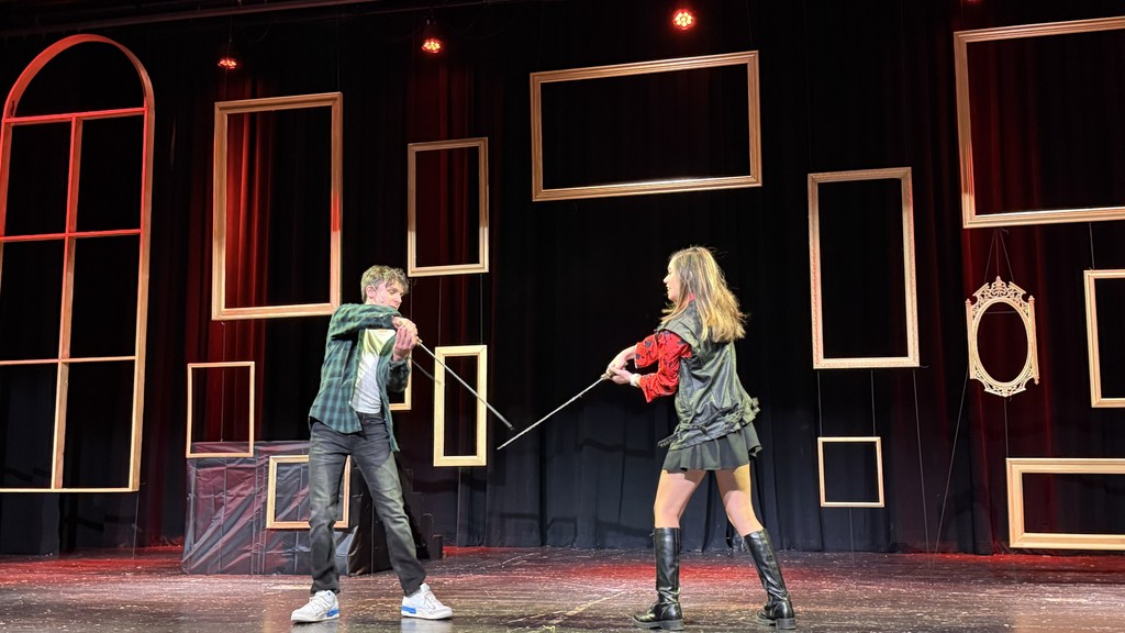 Two actors in a theater pose with swords, mid-fight. The stage backdrop features empty golden frames, creating a dramatic and intense atmosphere.