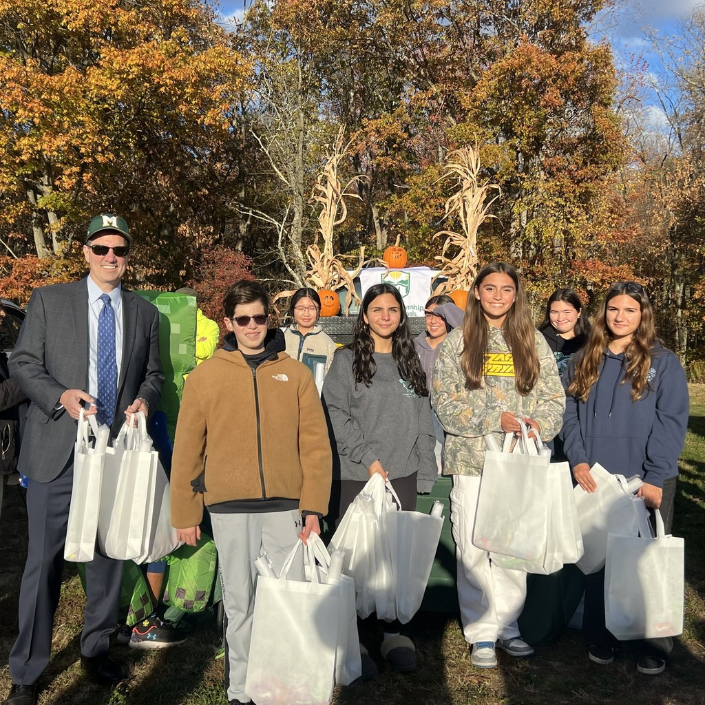 A group of nine people stand outside in a sunlit park, holding white bags. Behind them are autumn trees and Halloween decorations, including pumpkins. Everyone appears cheerful, creating a warm, community-oriented atmosphere.