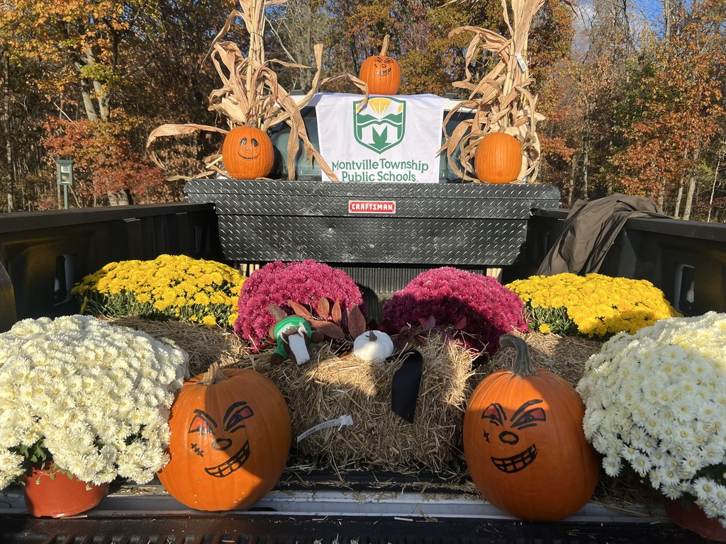 Festive truck bed with carved pumpkins, colorful chrysanthemums, corn stalks, and a "Montville Township Public Schools" banner, set against autumn trees.