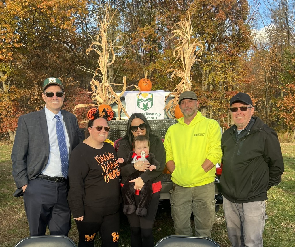 A group of six people poses outdoors with fall foliage and a banner behind them. One holds a baby dressed in a Halloween costume. Smiles and festive attire create a joyful, autumnal atmosphere.