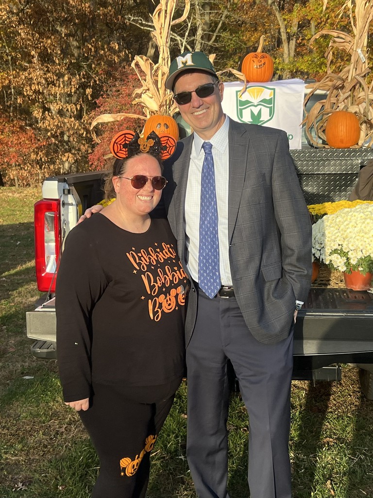 A smiling woman in Halloween attire stands beside a man in a suit and cap. They're in front of a truck with pumpkins and corn stalks, suggesting a festive fall setting.