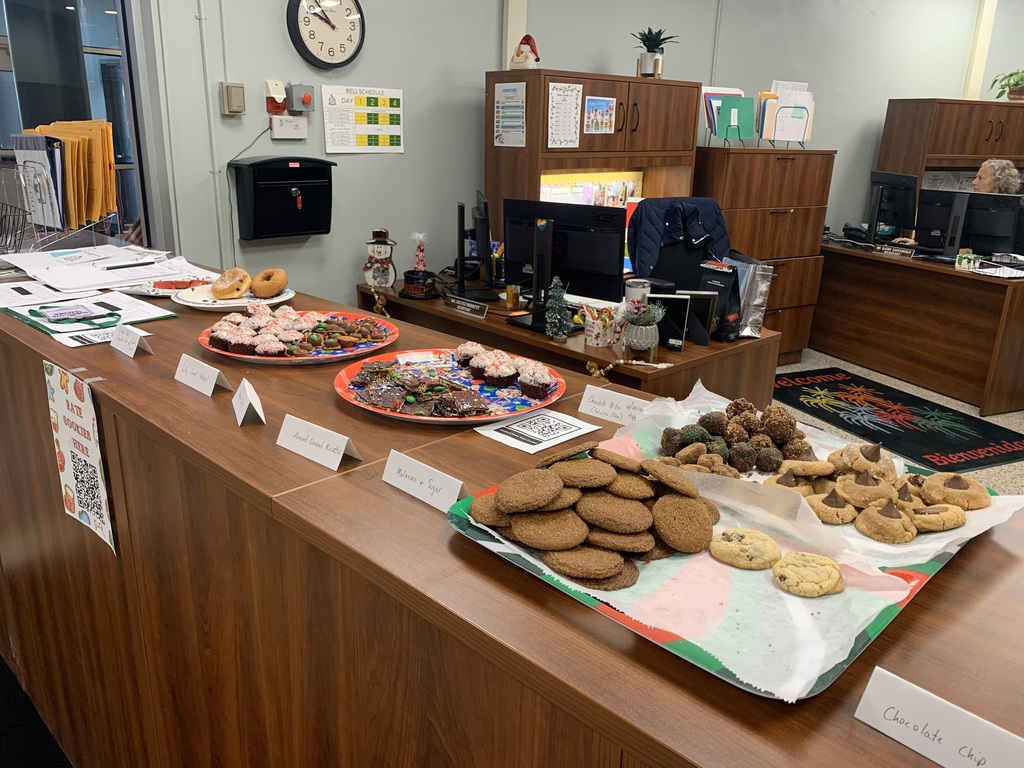 A table with a variety of holiday desserts, including donuts, cookies, and brownies, arranged on festive trays, with name cards. Cozy, inviting atmosphere.