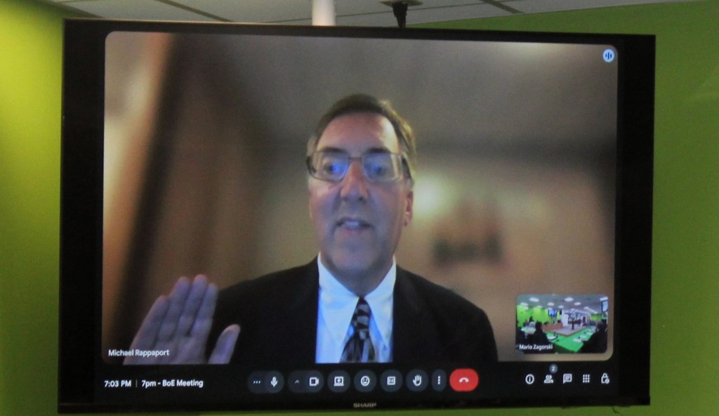 An individual in a suit and tie appears on a large screen during a virtual meeting. The person raises a hand, indicating communication or agreement. The background is blurred, focusing on the person speaking. Several video call icons are visible at the bottom of the screen.