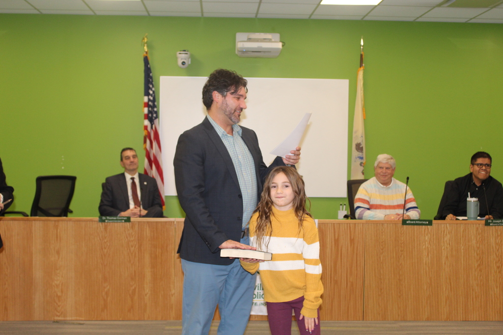 Man swearing in a young girl in a yellow and white sweater at a meeting. Officials sit at a table with green walls and flags in the background.