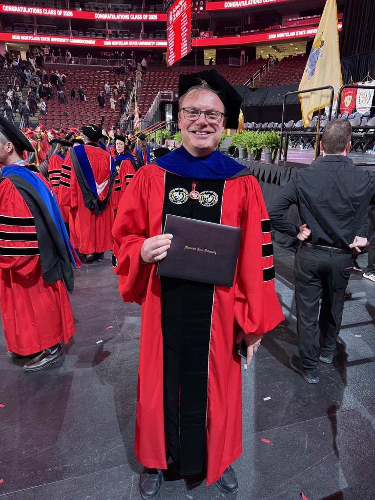 A graduate in a red gown and black cap smiles, holding a diploma at a commencement ceremony. Background shows fellow graduates and congratulatory banners.