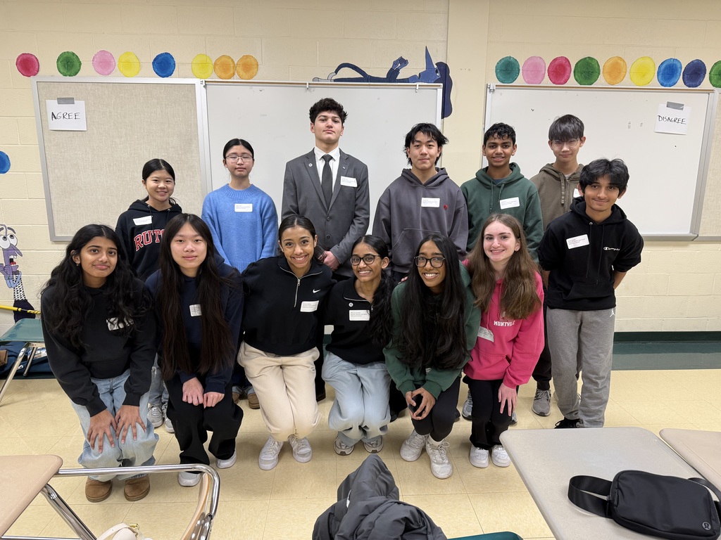 A diverse group of 13 students pose happily in a classroom. The back row stands, while the front row crouches. They wear casual clothes with name tags.