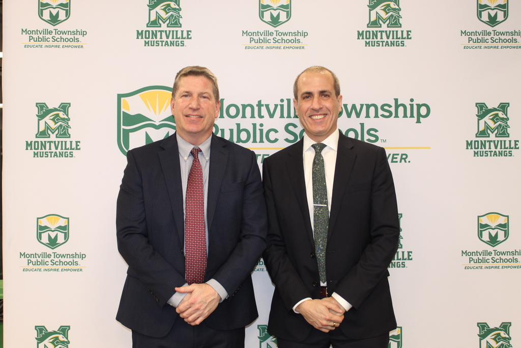 Two men in suits stand smiling in front of a Montville Township Public Schools backdrop. The mood is formal and professional.