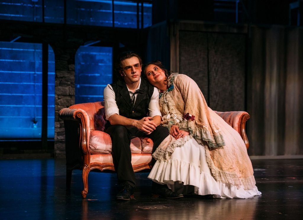 Two student actors perform a scene from Sweeney Todd while seated on a vintage-style couch on stage. One actor rests her head on the other’s shoulder as they wear period costumes, with cool blue lighting and an industrial-style set in the background.