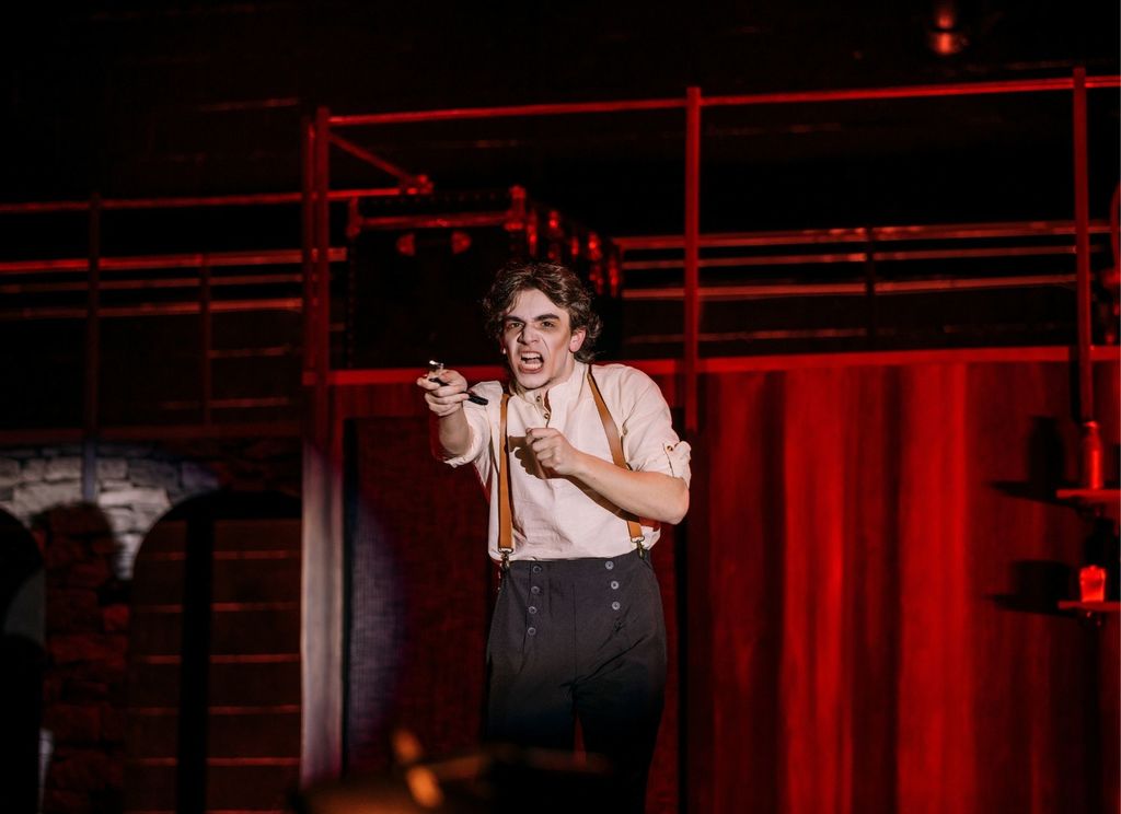 A student actor performs as Sweeney Todd on a dimly lit stage, dramatically pointing a straight razor toward the audience. The scene is bathed in red lighting, with an industrial-style set and railings in the background, creating a tense and dramatic atmosphere.