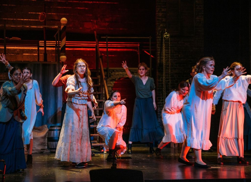 Students perform a dramatic scene from Sweeney Todd on stage, wearing period-style costumes and reaching their arms outward under red stage lighting. The set includes an industrial platform and barber pole, adding to the dark, theatrical atmosphere.