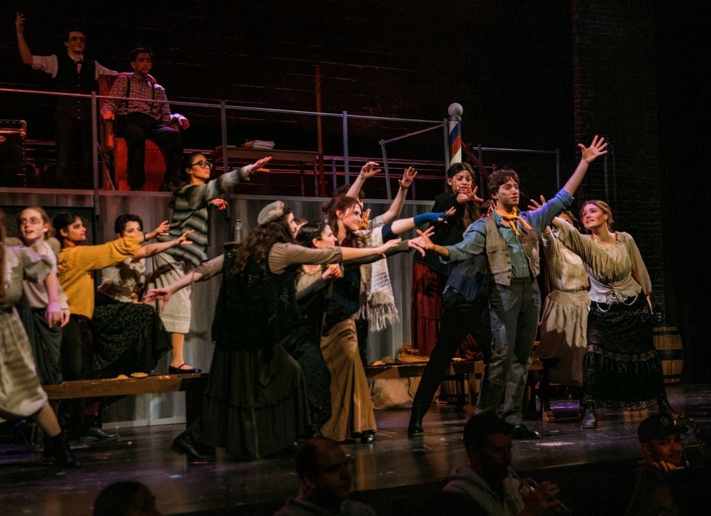 Students perform a scene from Sweeney Todd on stage, wearing period-style costumes and reaching their arms outward in a dramatic ensemble moment. The set features a barber chair and raised industrial platform, with warm stage lighting illuminating the large cast.