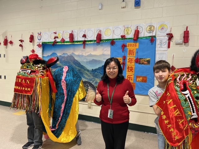 A woman in a red sweater stands smiling with thumbs up next to a child in colorful lion dance costumes. A festive backdrop features red decorations and a blue banner with Chinese characters, creating a joyful atmosphere.