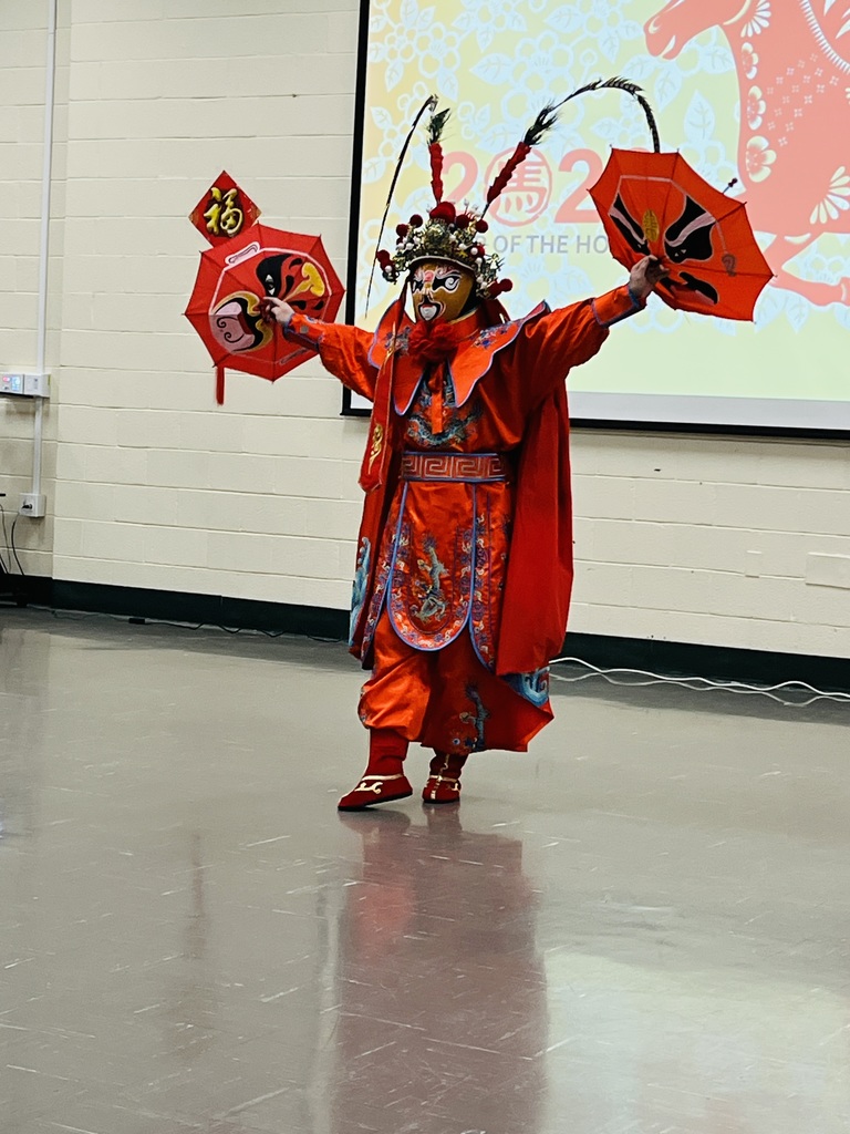 A performer in vibrant traditional Chinese costume and mask, adorned with elaborate headgear, stands confidently with outstretched arms holding fans.