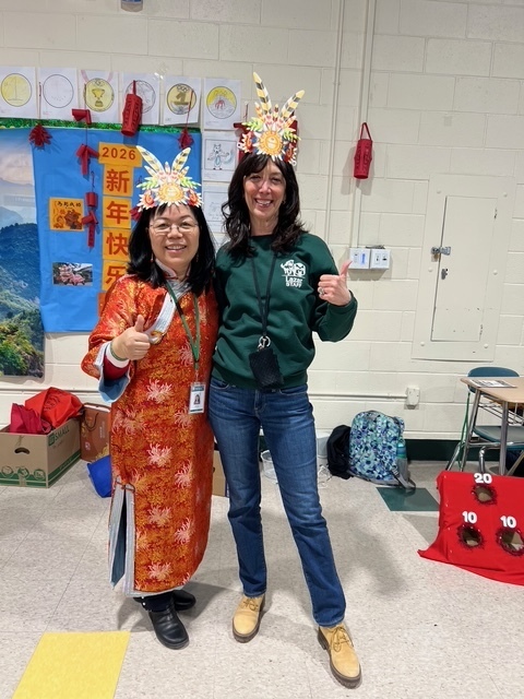 Two women smiling and giving thumbs up at a festive event. Both wear colorful rooster-themed headbands. A bright, decorative banner is in the background.