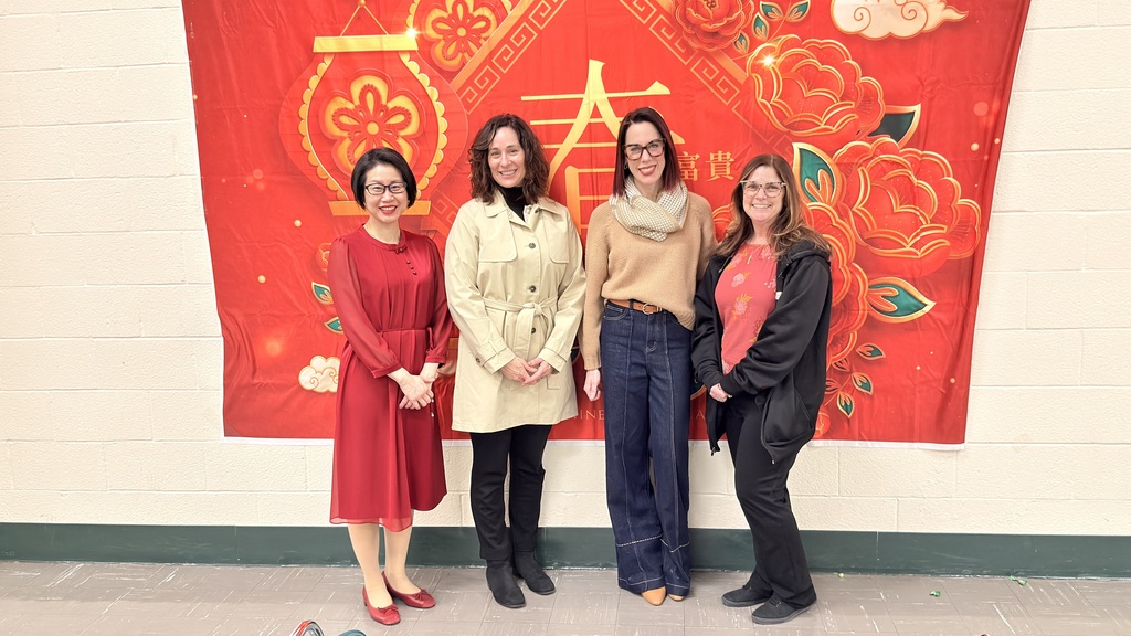 Four women stand smiling in front of a red Chinese New Year-themed backdrop with floral and geometric designs. The setting is festive and warm.