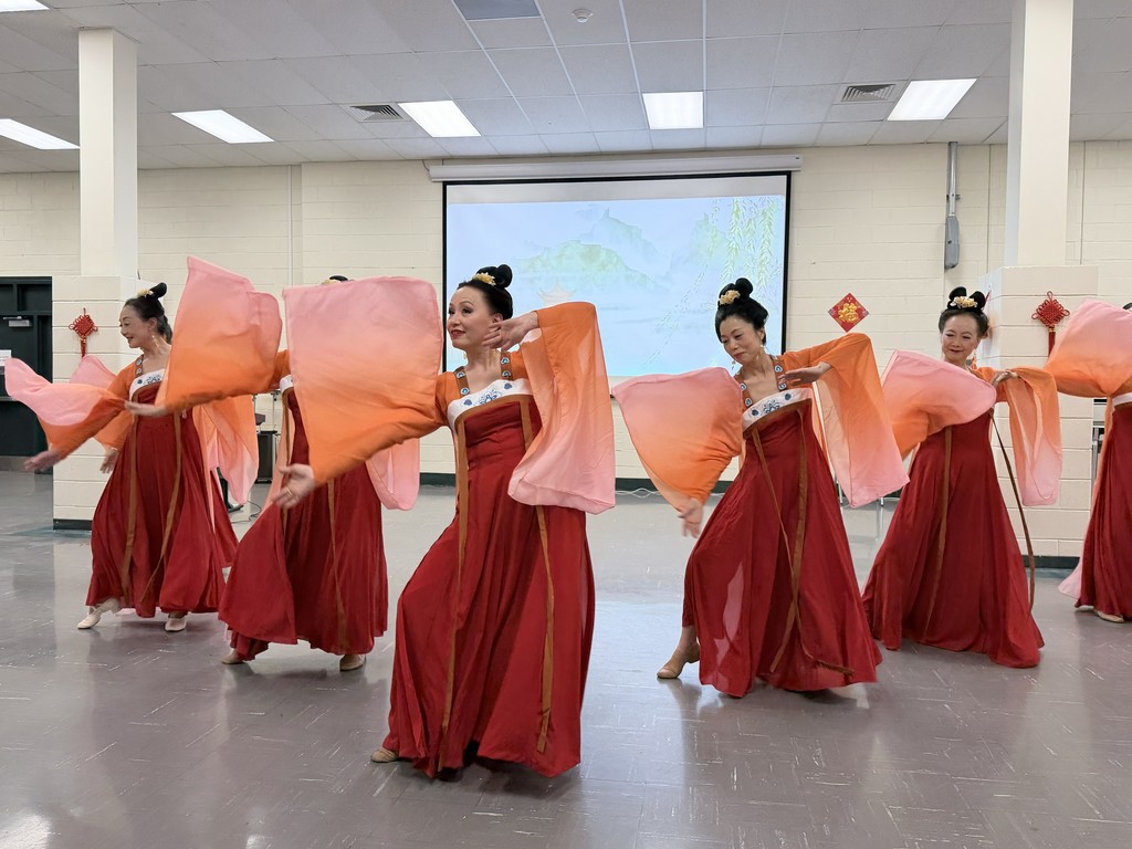 A group of dancers in red and pink traditional attire perform gracefully, their flowing sleeves creating dynamic motion. A serene backdrop enhances the scene.