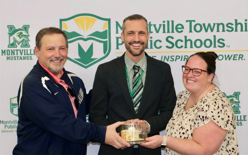 Three people smiling in front of a Montville Township Public Schools banner. The person in the center holds a small cake. The atmosphere is celebratory.