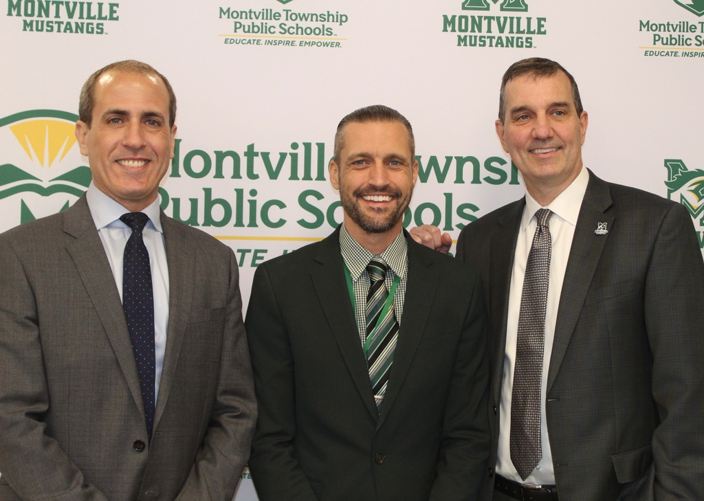 Three men in suits stand smiling in front of a backdrop with "Montville Township Public Schools" and "Montville Mustangs" logos, exuding a formal, friendly tone.