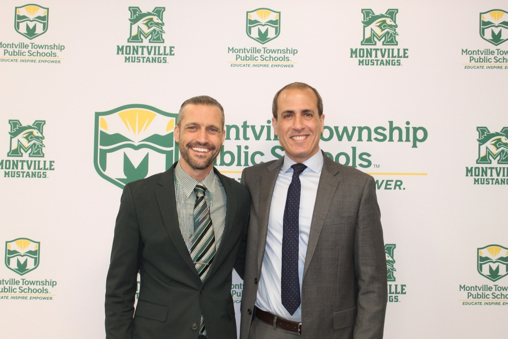 Two men in suits smiling warmly, standing in front of a Montville Township Public Schools backdrop, conveying a professional and welcoming atmosphere.