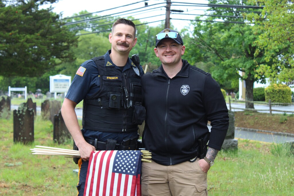 Two smiling police officers stand together outdoors. One holds several flags, including an American flag. They are in a cemetery with trees in the background.